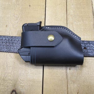 May include: A black leather holster with a gun inside, attached to a textured black belt. The holster has white stitching and a brass-colored snap closure. The belt is secured with a buckle.