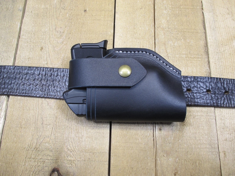 May include: A black leather holster with a gun inside, attached to a textured black belt. The holster has white stitching and a brass-colored snap closure. The belt is secured with a buckle.