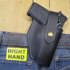 May include: Black leather holster with a handgun, attached to blue denim jeans. The holster is secured with a brass-coloured button. A yellow tag with the words "RIGHT HAND" is visible.