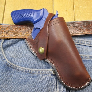 May include: A brown leather holster with a brass snap closure containing a blue revolver. The holster is attached to a brown leather belt with an embossed floral pattern, which is worn over a pair of blue denim jeans.