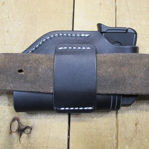 May include: Black leather holster with white stitching, designed to be worn on a belt. The holster is holding a black handgun. The belt is brown and has several holes.