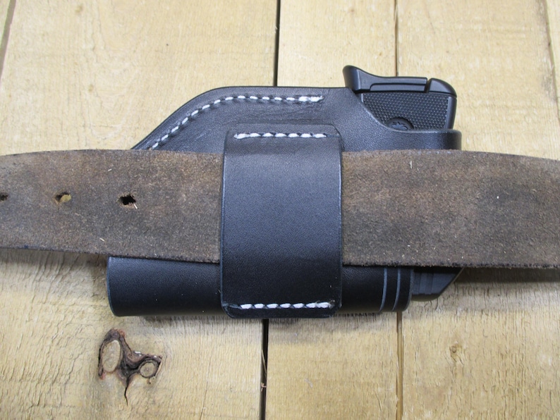 May include: Black leather holster with white stitching, designed to be worn on a belt. The holster is holding a black handgun. The belt is brown and has several holes.
