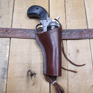 May include: A brown leather holster with a silver and black revolver inside. The holster is attached to a brown leather belt with an embossed floral design.
