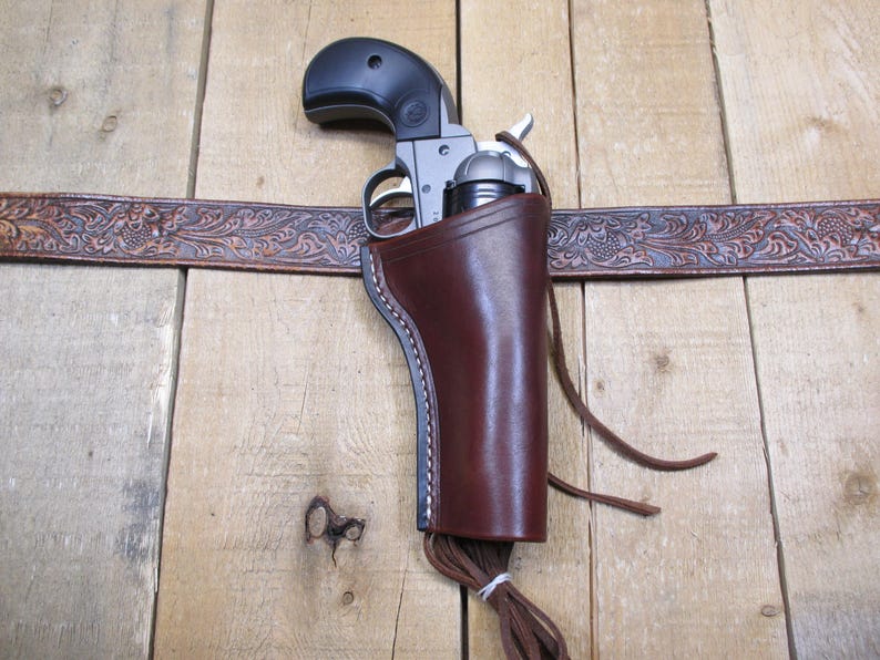 May include: A brown leather holster with a silver and black revolver inside. The holster is attached to a brown leather belt with an embossed floral design.