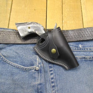Peut inclure: Un étui en cuir noir avec un revolver argenté, attaché à une ceinture grise sur un jean bleu. L'étui a une fermeture à bouton et des coutures blanches. La poignée du revolver a un motif.
