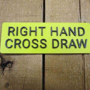 May include: A rectangular, neon yellow sign with the words "RIGHT HAND CROSS DRAW" in black, raised lettering. The sign is set against a wooden background.