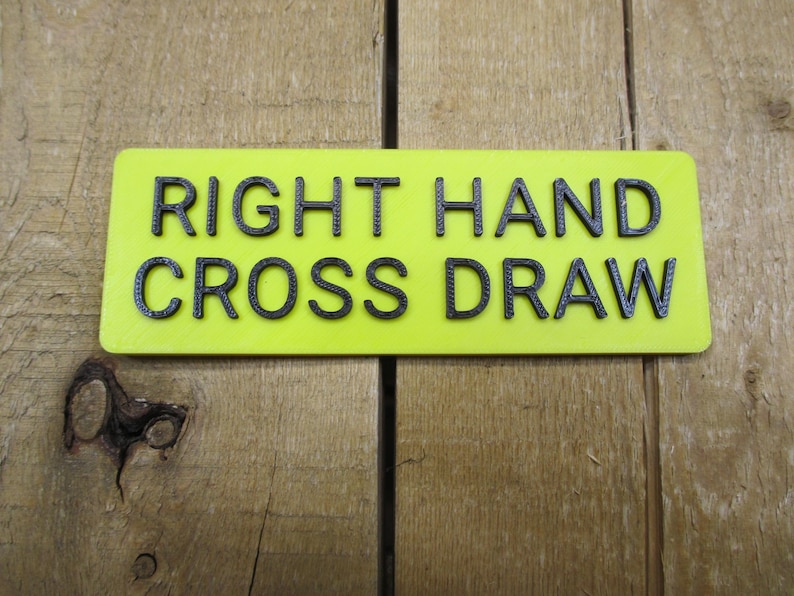 May include: A rectangular, neon yellow sign with the words "RIGHT HAND CROSS DRAW" in black, raised lettering. The sign is set against a wooden background.