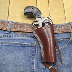 May include: A brown leather holster with a fringe detail, holding a silver revolver with a black handle. The holster is attached to a brown leather belt with an intricate floral design, worn over blue jeans.