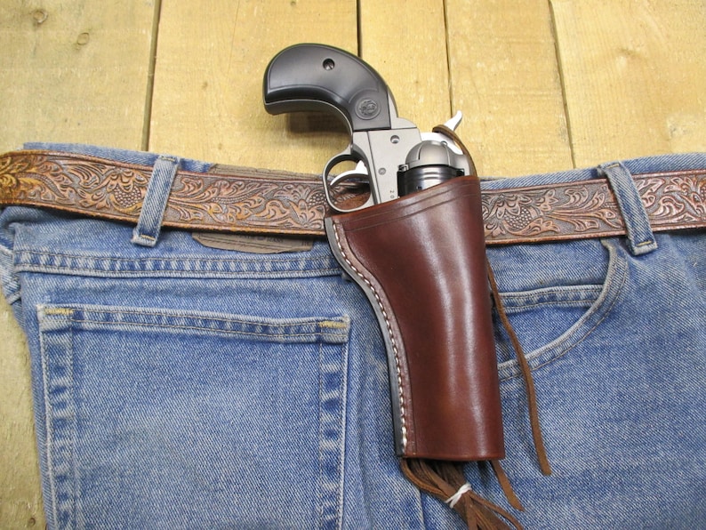 May include: A brown leather holster with a fringe detail, holding a silver revolver with a black handle. The holster is attached to a brown leather belt with an intricate floral design, worn over blue jeans.