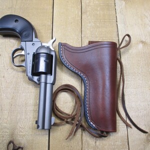 May include: A silver revolver with a black grip and a brown leather holster with white stitching. The holster has a loop of brown leather cord. The gun is lying on a wooden surface.