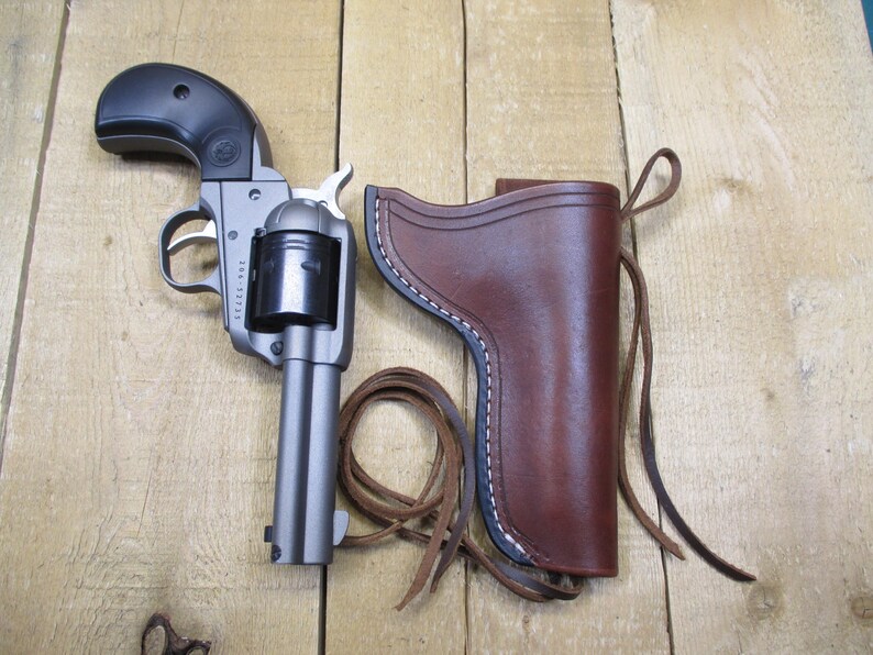 May include: A silver revolver with a black grip and a brown leather holster with white stitching. The holster has a loop of brown leather cord. The gun is lying on a wooden surface.