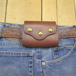 Op de afbeelding: Een bruine leren riem met een gegraveerd ontwerp en een bruine leren tas met drie messing studs. De tas is bevestigd aan de riem en wordt gedragen over een blauwe jeans.