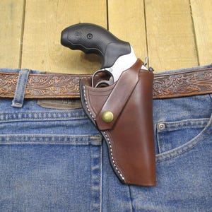 May include: A brown leather holster with a silver revolver inside. The holster is attached to a brown leather belt with an embossed floral design. The belt is worn over a pair of blue jeans.