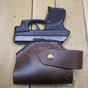 May include: A black handgun rests atop a brown leather holster. The holster features decorative stitching and a snap closure. The gun is marked "Ruger Prescott - AZ - USA".
