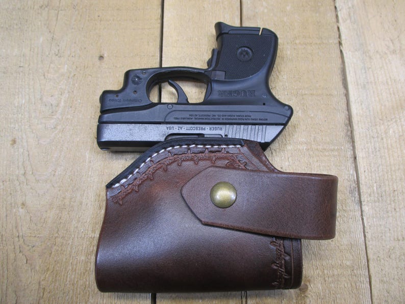 May include: A black handgun rests atop a brown leather holster. The holster features decorative stitching and a snap closure. The gun is marked "Ruger Prescott - AZ - USA".