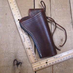 May include: A brown leather holster with white stitching and a leather tie. The holster is shown with two wooden rulers, one measuring up to 30 inches. The holster is on a wooden surface.