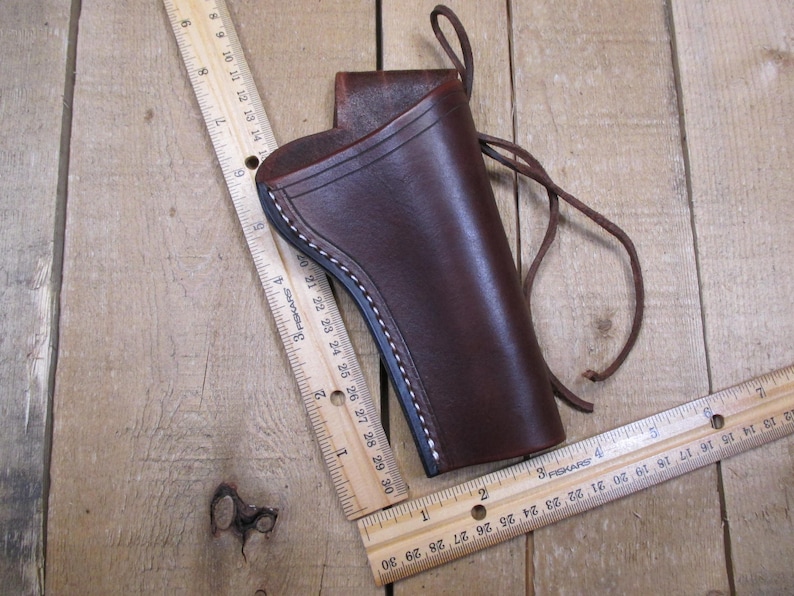 May include: A brown leather holster with white stitching and a leather tie. The holster is shown with two wooden rulers, one measuring up to 30 inches. The holster is on a wooden surface.