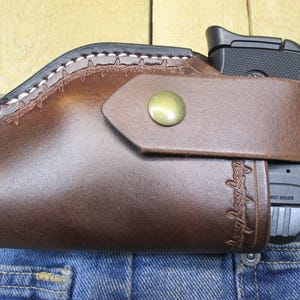 May include: A brown leather holster with a black handgun. The holster is attached to a blue denim jean. The holster has a decorative stitched design and a brass button closure. The holster is designed to be worn on a belt.