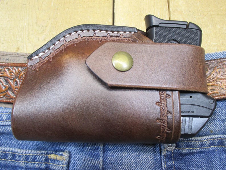 May include: A brown leather holster with a black handgun. The holster is attached to a blue denim jean. The holster has a decorative stitched design and a brass button closure. The holster is designed to be worn on a belt.
