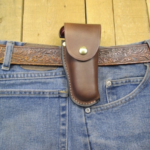 May include: A brown leather belt with an intricate floral design is worn over a pair of blue jeans. A brown leather holster is attached to the belt and is positioned over the right front pocket of the jeans.