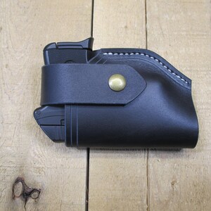 May include: Black leather holster with a gun inside. The holster has a strap with a brass button closure. White stitching accents the top edge. The gun is partially visible.