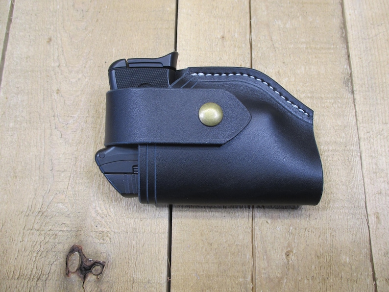 May include: Black leather holster with a gun inside. The holster has a strap with a brass button closure. White stitching accents the top edge. The gun is partially visible.