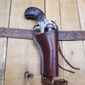 May include: A brown leather holster with a silver revolver inside. The holster is attached to a brown leather belt with an embossed floral design.