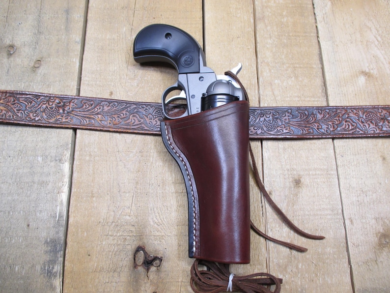 May include: A brown leather holster with a silver revolver inside. The holster is attached to a brown leather belt with an embossed floral design.