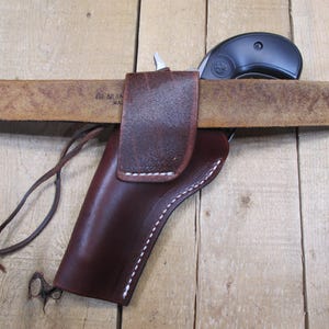 May include: A brown leather holster with white stitching, designed to be worn on a belt. A black revolver is partially visible, resting in the holster. The belt is a light brown color and the background is a wooden surface.