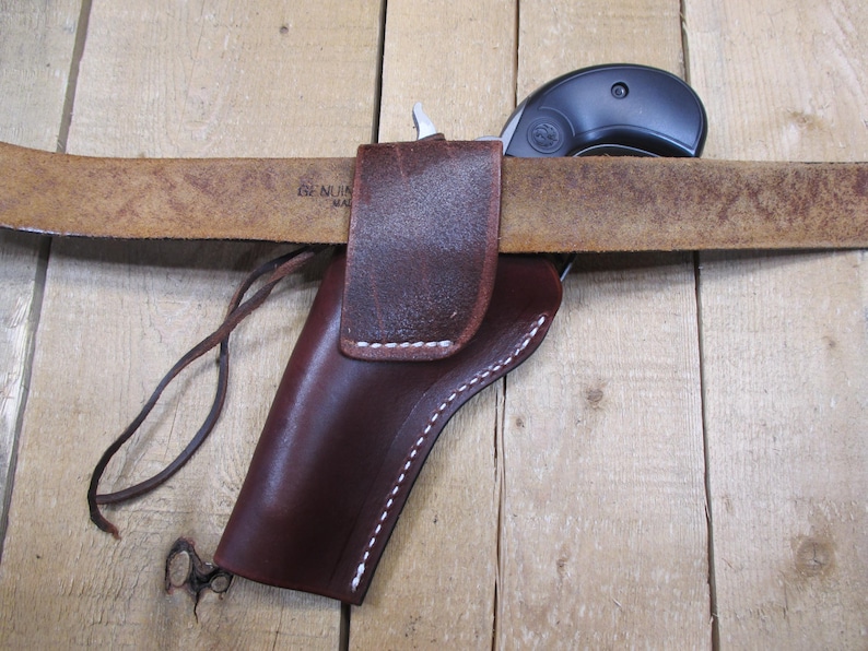 May include: A brown leather holster with white stitching, designed to be worn on a belt. A black revolver is partially visible, resting in the holster. The belt is a light brown color and the background is a wooden surface.