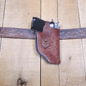 May include: A brown leather holster with a black handgun, secured on a decorative brown leather belt. The holster has a textured surface and a flap with a brass button. The belt features an embossed floral pattern.