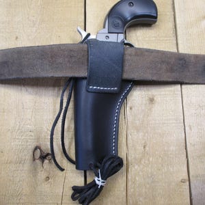 以下が含まれることがあります： 銃器を収納するために設計された、白いステッチのディテールが施された黒い革製のホルスター。ホルスターは茶色の革製のベルトに取り付けられています。