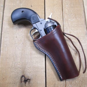 May include: A silver and black revolver is partially holstered in a brown leather holster. The holster has white stitching and a leather tie. The revolver is resting on a wooden surface.