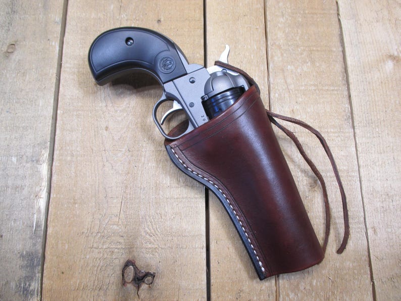 May include: A silver and black revolver is partially holstered in a brown leather holster. The holster has white stitching and a leather tie. The revolver is resting on a wooden surface.