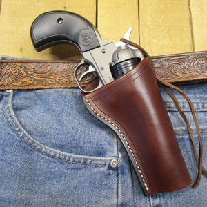 May include: A silver and black revolver is holstered in a brown leather holster, secured to a pair of blue denim jeans. A brown leather belt with an embossed floral design is visible above the jeans. The holster is stitched with white thread.
