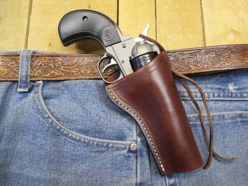 May include: A silver and black revolver is holstered in a brown leather holster, secured to a pair of blue denim jeans. A brown leather belt with an embossed floral design is visible above the jeans. The holster is stitched with white thread.