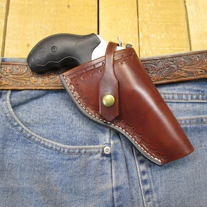 Può includere: Una fondina in pelle marrone con un bottone color oro, che contiene un revolver nero e argento. La fondina è attaccata a una cintura marrone fantasia, indossata con jeans blu.