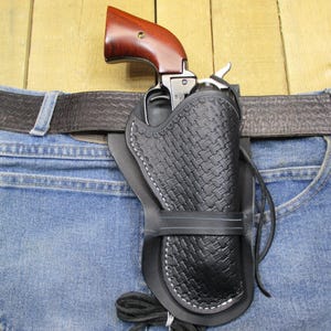Puede incluir: Una funda de cuero negro con patrón de tejido de cesta y costuras blancas, que contiene un revólver con armazón de metal oscuro y empuñadura de madera de color marrón rojizo pulido, sujeta a un cinturón de cuero marrón sobre tela vaquera.