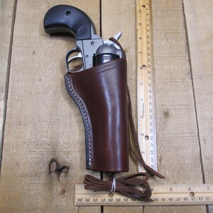 May include: A brown leather holster with a stitched edge, designed to hold a small silver revolver. The holster has a strap and a loop for attaching to a belt.