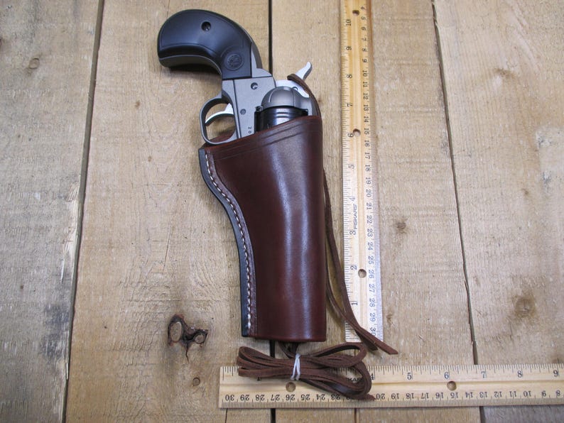 May include: A brown leather holster with a stitched edge, designed to hold a small silver revolver. The holster has a strap and a loop for attaching to a belt.