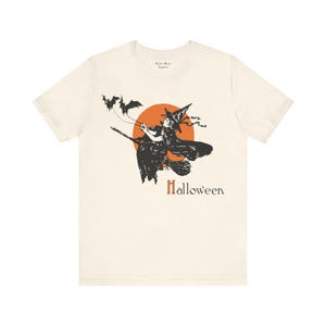 Può includere: T-shirt color crema con grafica a tema Halloween. Il design presenta una strega nera su una scopa, un sole arancione e la scritta "Halloween" in marrone. È un capo d'abbigliamento casual.