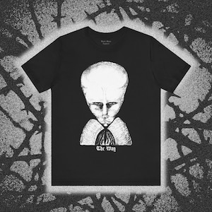 May include: A black short-sleeve t-shirt featuring a white graphic illustration of a large-headed figure with a somber expression. Below the figure, the text "The Way" is printed in a gothic-style font. The shirt has a crew neckline.