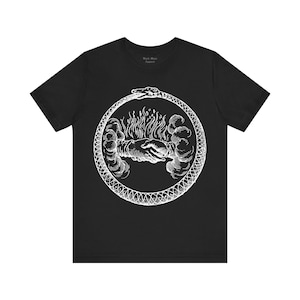 以下が含まれることがあります： 黒いTシャツ。白いウロボロス（自らを食べる蛇）のデザインで、炎を握る手を囲んでいます。「Black Mass Apparel」の文字が上部にプリントされています。