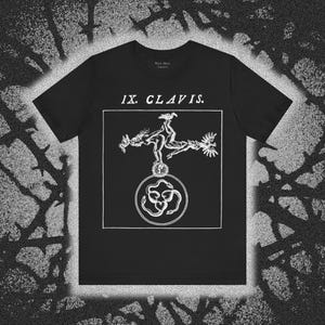 May include: A black short-sleeve t-shirt featuring a white graphic print. The design includes the text IX. CLAVIS. above a square frame containing two figures balancing on a circle, which encloses three intertwined snakes forming a heart shape.