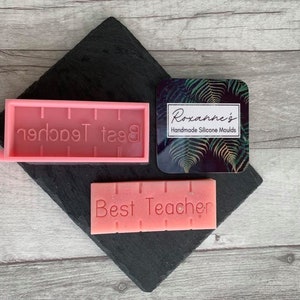 May include: Pink silicone mold with the words "Best Teacher" inscribed. A pink bar of soap with the same inscription is next to the mold. A black square with a tropical leaf design and the text "Roxanne's Handmade Silicone Molds" is in the upper right corner.