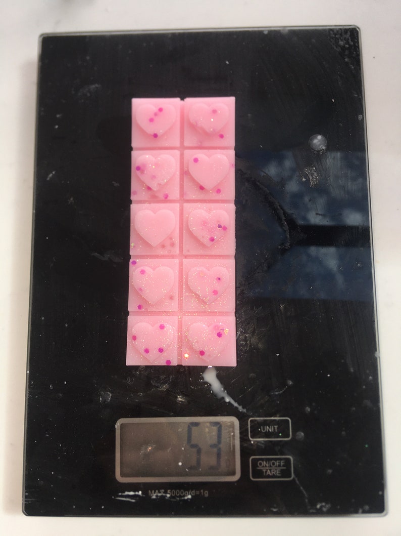 May include: A pink wax melt with a heart design and pink glitter. The wax melt is on a digital scale that reads 53 grams.