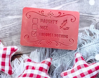 May include: Red Christmas card with a checklist that says "Naughty", "Nice", and "I Regret Nothing". The "I Regret Nothing" box is checked. The card is on a white and gray fuzzy surface with red and white gingham stars.