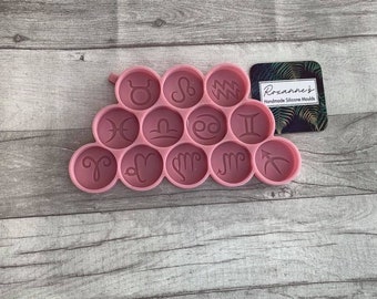 May include: Pink silicone mold with 12 circular impressions, each featuring a different zodiac sign. The mold is shaped like a pyramid with a rounded top. The text "Roxanne's Handmade Silicone Moulds" is printed on a small card with a green and brown tropical leaf design.