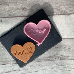 May include: A pink heart-shaped silicone mold with the word "mum" written inside. A brown heart-shaped cookie with the word "mum" written inside is next to the mold.