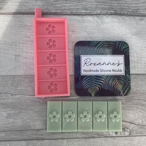 Op de afbeelding: Een roze siliconen mal met vijf bloemvormige holtes. De mal ligt naast een groene wasbalk met drie bloemvormige indrukken. Een zwart-groene kaart met de tekst "Roxanne's Handmade Silicone Moulds" is op de achtergrond.